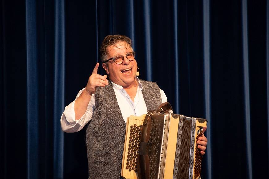 Schösu gibt bei uns auf der Bühne, vor dem dunkelblauen Bühnenvorhang mit seiner Handharmonika einen Witz zum Besten.
